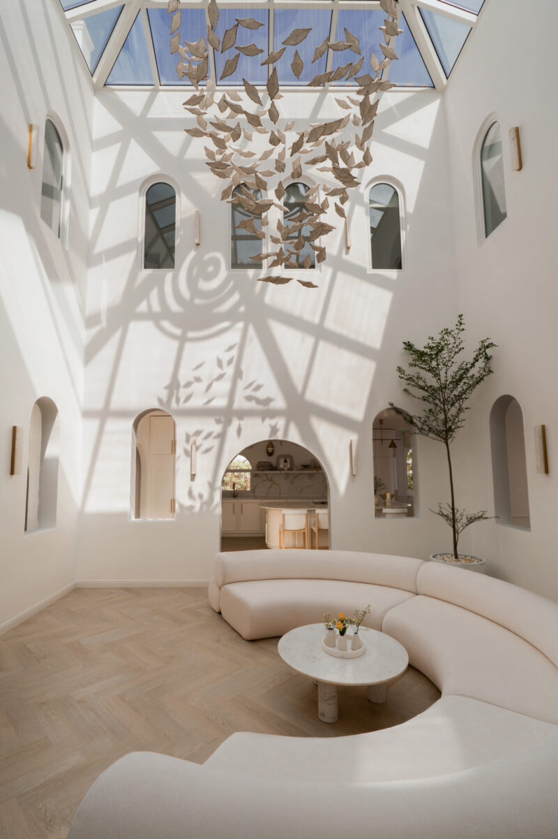 A bright, modern atrium by D’Ora Tokai Designs features white curved seating, a round table, skylight, abstract hanging sculpture, large windows, and a potted tree on a light wood herringbone floor.