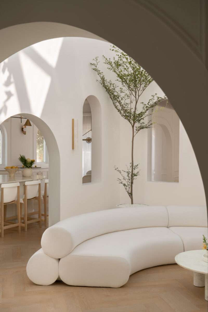 Minimalist living room with curved white sofa, a round coffee table, arched doorways, indoor tree, and light wood floor. Sunlight casts shadows on white walls.