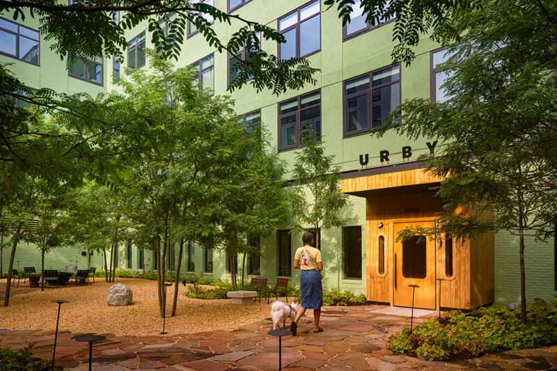 A person stands with a dog in a landscaped courtyard outside Fishtown Urby, a green apartment building with a wooden entrance labeled 