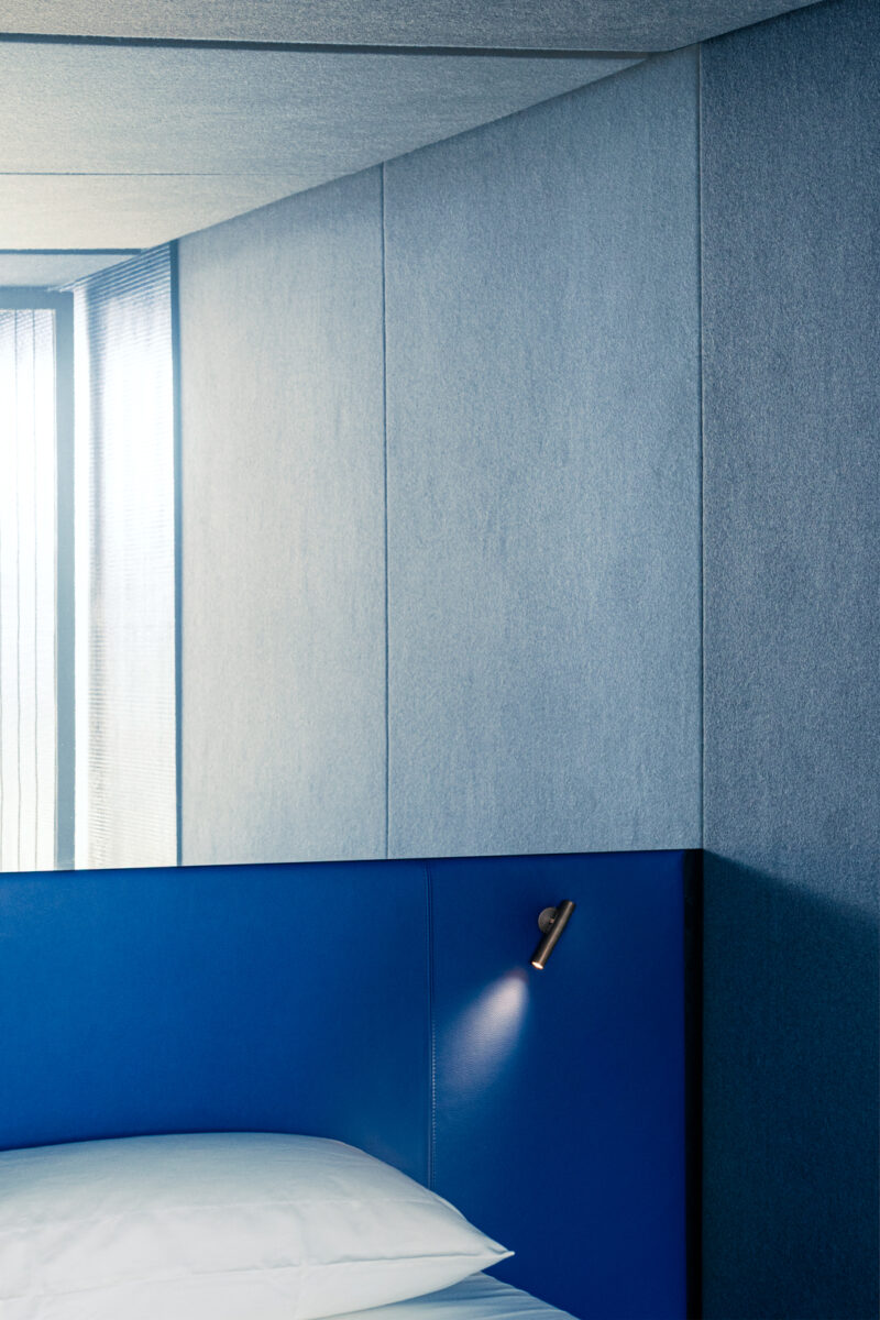 A modern bedroom with blue upholstered walls, a blue headboard, a pillow, and an angled reading light—this serene space feels like an m2atelier Crafted Capsule beside a window with sheer curtains.