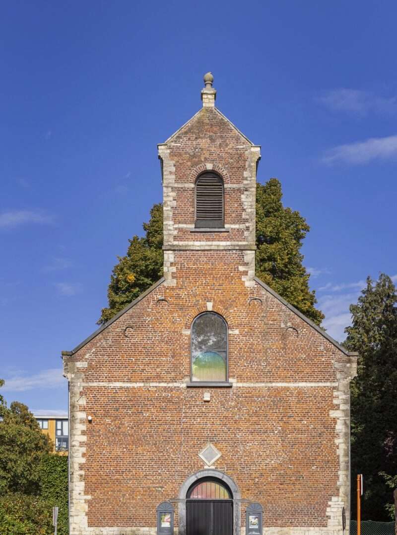 A small brick church with a central arched window and bell tower stands in front of trees, with a blue and yellow barrier at its base under a clear blue sky.