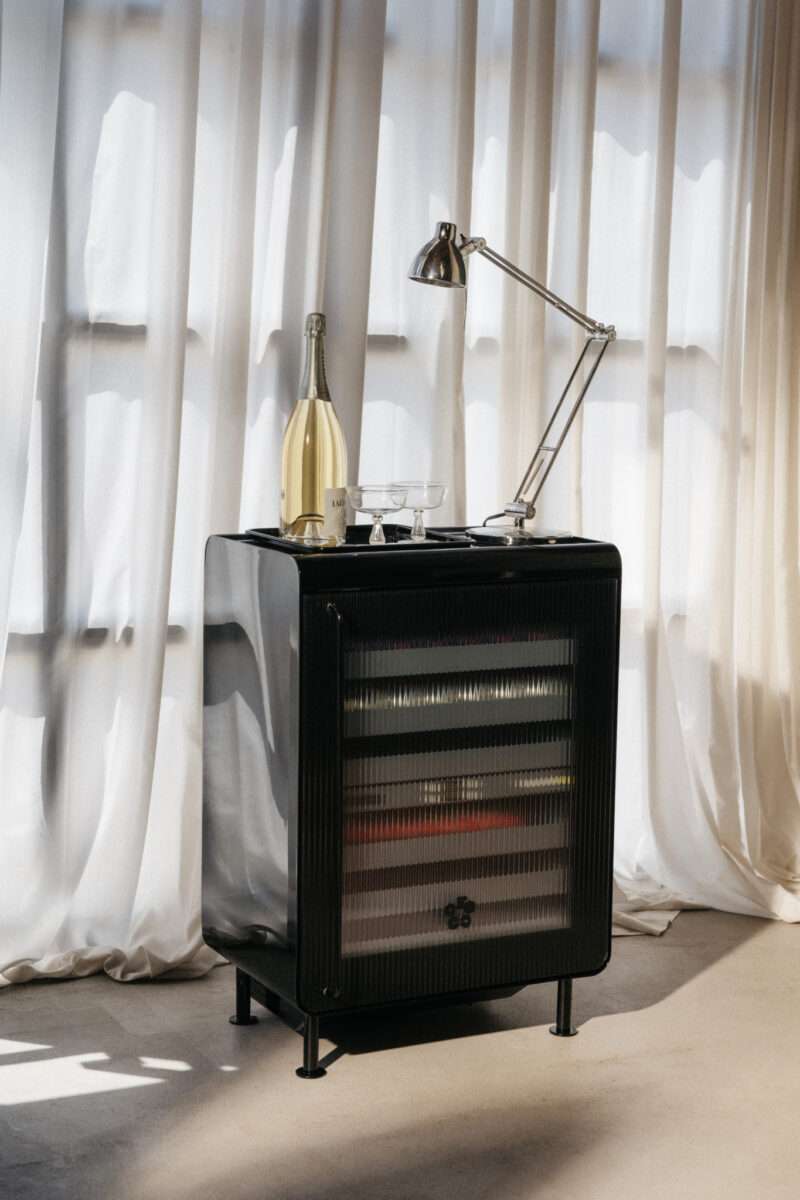 A black mini fridge with a glass door stands in front of sheer curtains, topped with a lamp, a bottle of white wine, and two glasses.