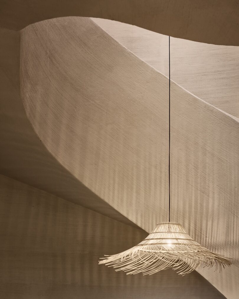 A woven pendant light hangs from the ceiling, casting shadows on curved, textured beige walls in a minimalist interior space.