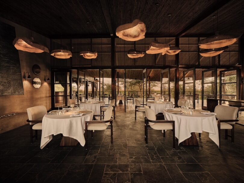 Elegant restaurant interior with round tables covered in white cloths, cushioned chairs, large windows, and warm ceiling lights.