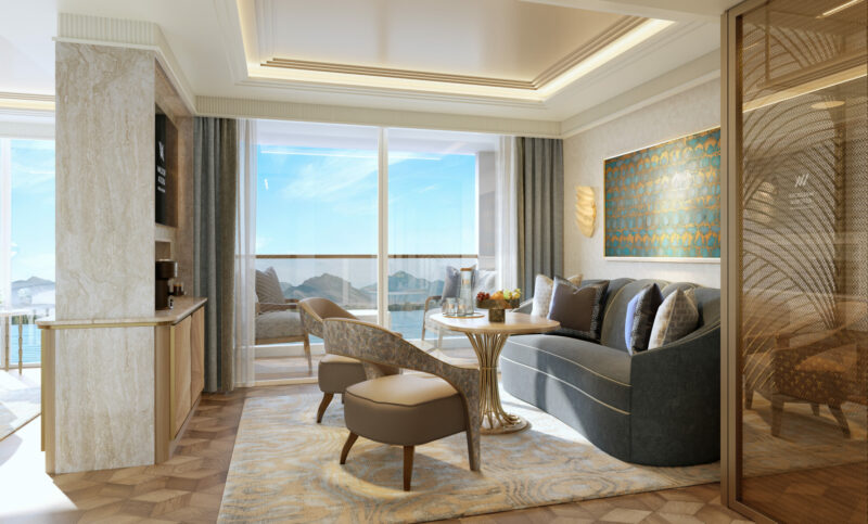 Modern hotel suite with seating area, artwork, and balcony overlooking water and mountains, featuring neutral tones and floor-to-ceiling windows.