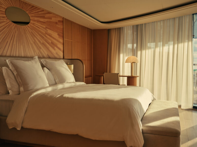 Modern hotel room with a large neatly made bed, cushioned bench, wood paneling, sunburst wall art, desk, lamp, and sheer curtains letting in natural daylight.