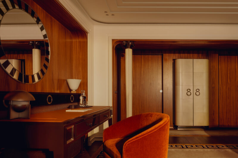A modern hotel room features a wooden desk with a lamp, an orange chair, a large mirror, and a wardrobe with decorative door handles.