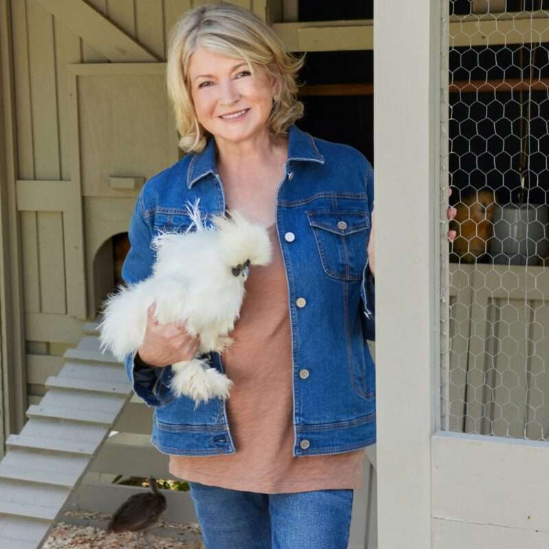 Woman wearing a denim jacket stands by a chicken coop, holding a fluffy white chicken and smiling at the camera, channeling Martha Stewart's signature blend of warmth and style.