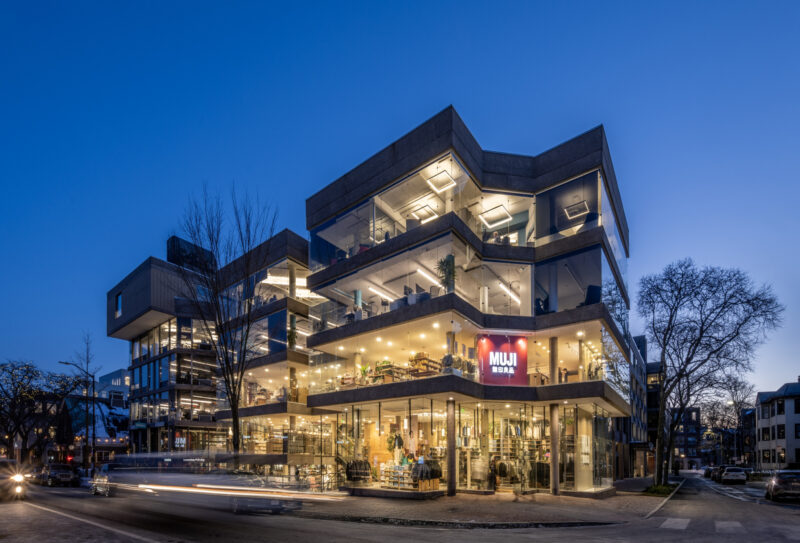 A modern, multi-story building at dusk features large glass windows and illuminated interiors. A red 