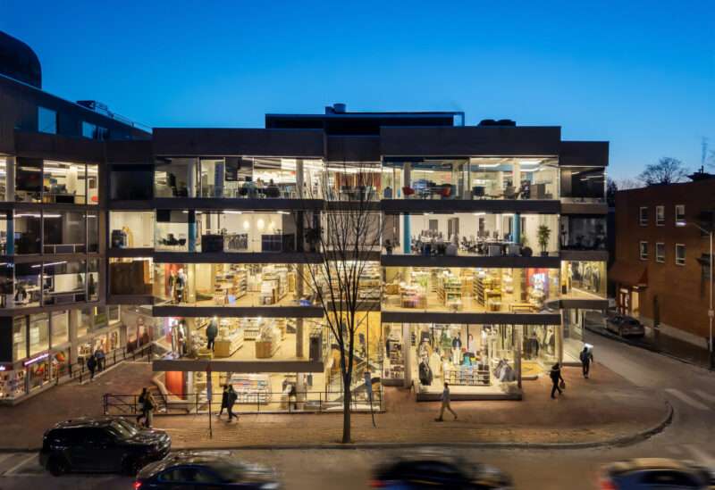 A modern, glass-walled building at dusk shows three brightly lit floors with people inside and outside, as cars and pedestrians move along the street in the foreground near MUJI Harvard Square.