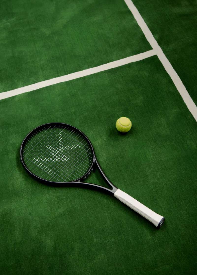 A black tennis racket and a yellow tennis ball sit on a green tennis court with white boundary lines, evoking the crisp, contemporary elegance found in the Nordic Knots Grand Open rug collection.