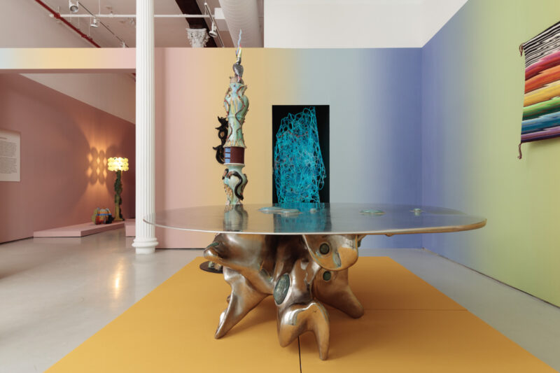 Art gallery space with a round table featuring an organic, sculptural base, abstract sculpture on top, a blue wire artwork, and a colorful textile on pastel gradient walls.