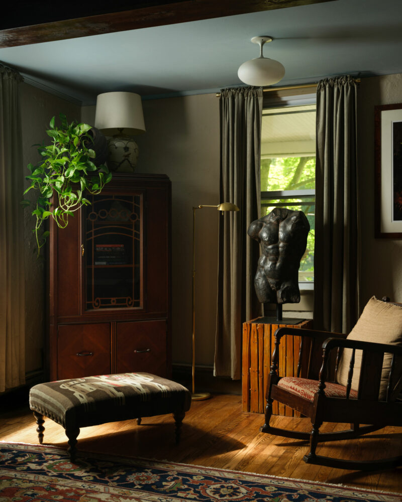 A cozy living room corner with a wooden cabinet, potted plant, floor lamp, cushioned chair, patterned ottoman, sculpture on a crate, and a window with curtains letting in natural light.