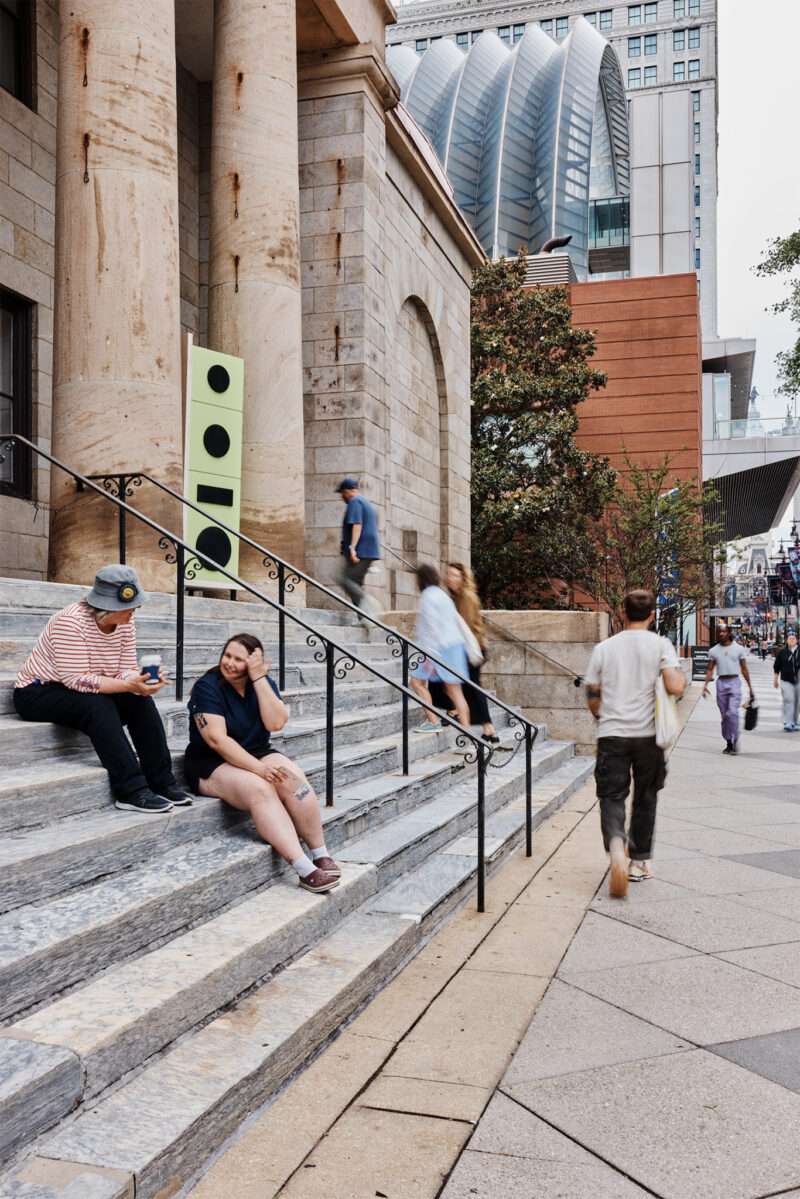 People sit and talk on stone steps outside a large building, while others walk along the sidewalk; modern buildings are visible in the background.