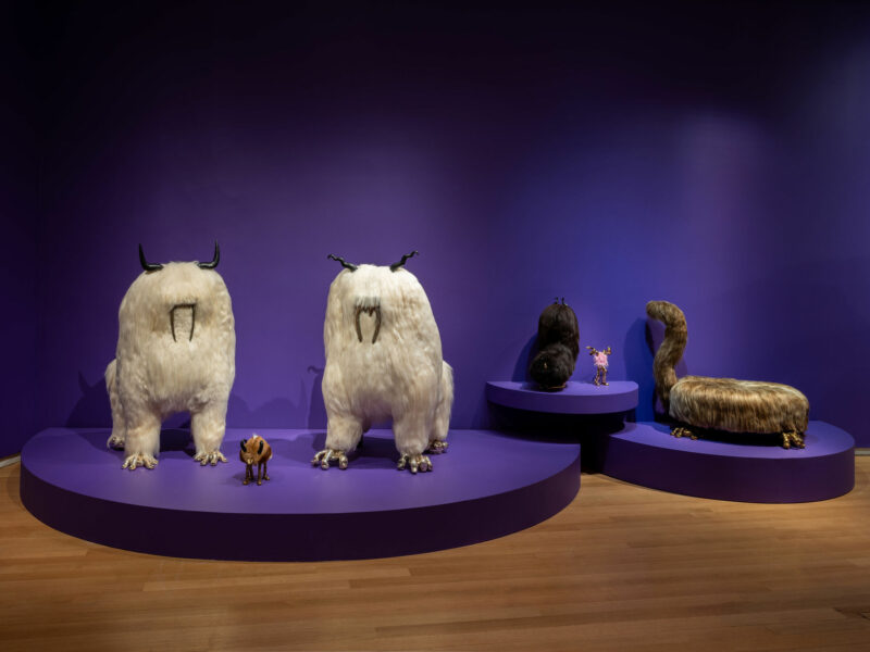 Five large, fantastical animal sculptures with exaggerated features and fur—characteristic of the Haas Brothers' Uncanny Valley aesthetic—are displayed on purple platforms against a purple wall in a gallery setting.