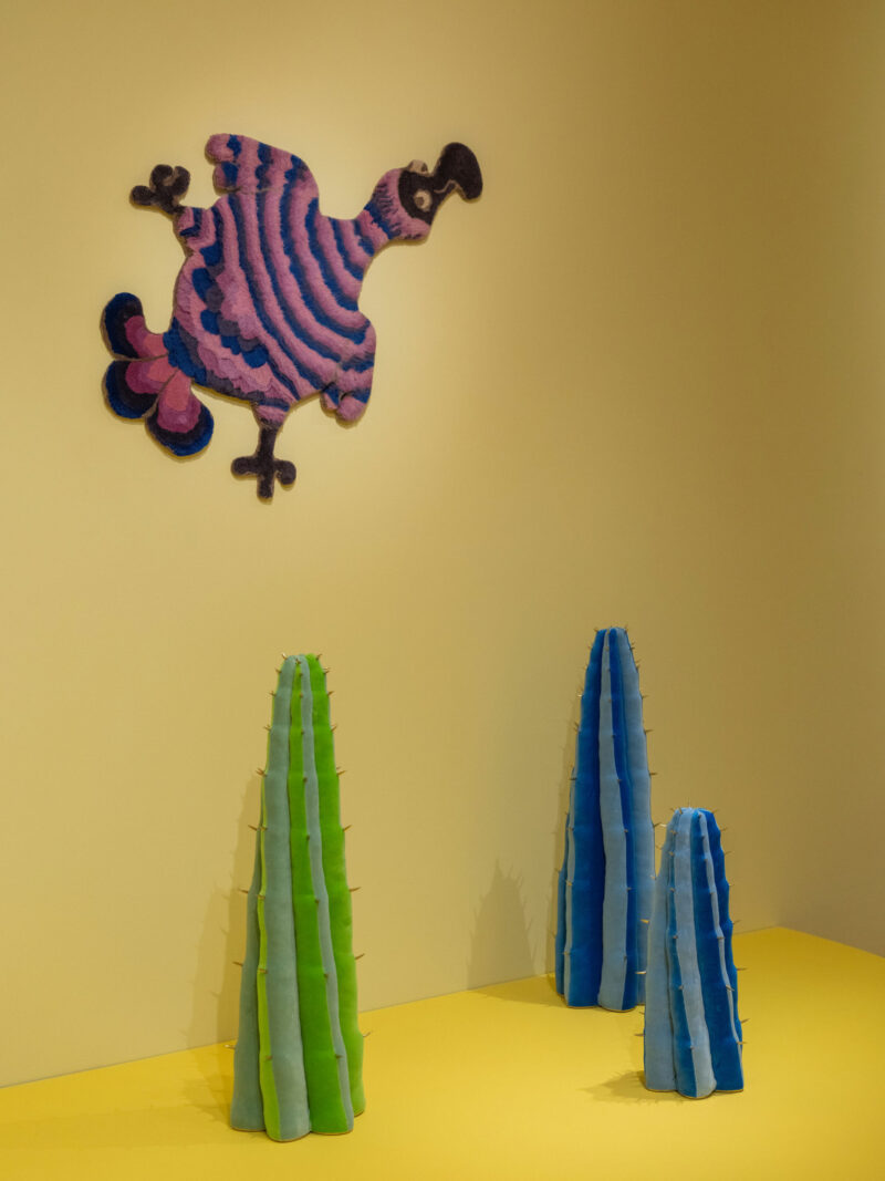 A colorful striped bird art piece hangs on a yellow wall above three vertical Haas Brothers Uncanny Valley-inspired cactus sculptures in green and blue hues on a yellow floor.