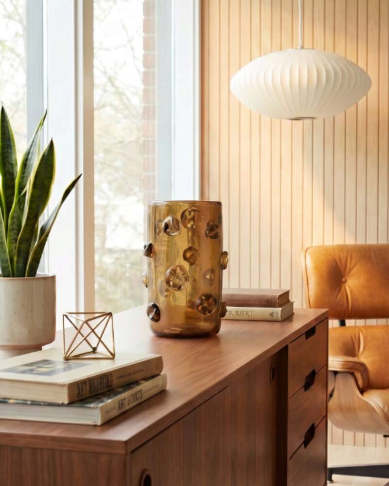 A modern office with a wooden desk, brown leather chair, potted plant, books, amber vase, and a white pendant lamp—enhanced by subtle touches of Modern African Decor—set against vertical wood paneling and large windows.