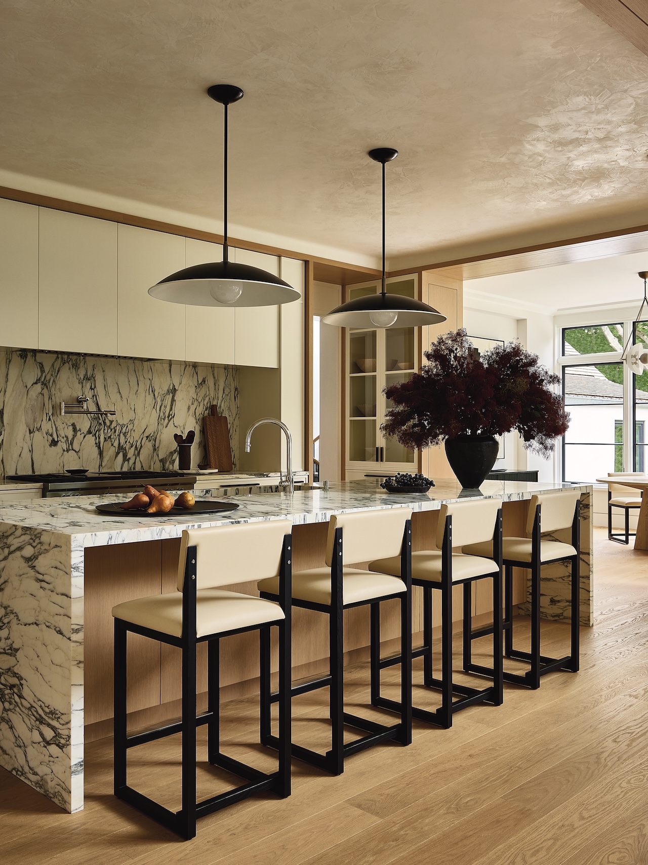 Modern kitchen with a marble island, four bar stools, pendant lights, light wood floors, and a large vase with dark red foliage.