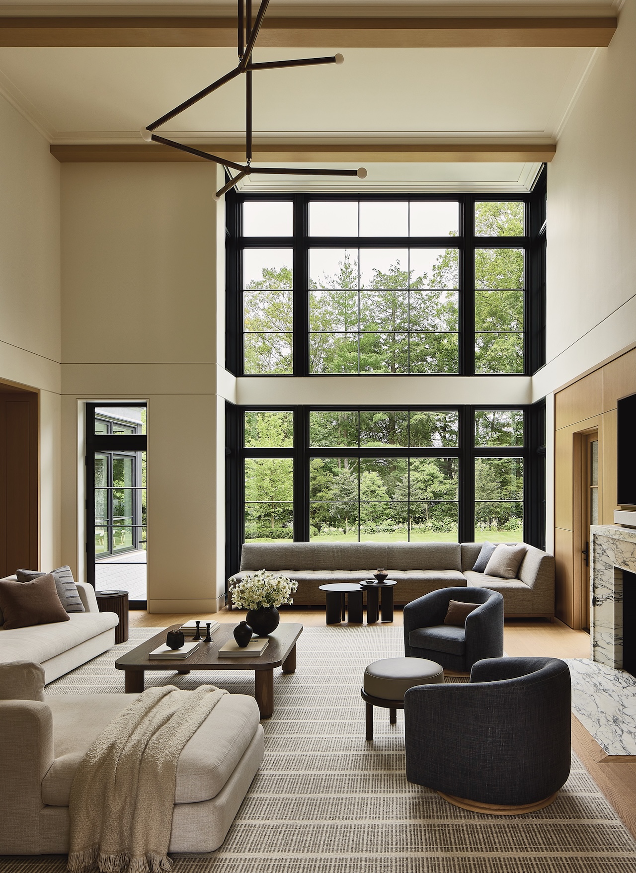 Modern living room with large floor-to-ceiling windows, neutral furniture, a marble fireplace, and a view of green trees outside.