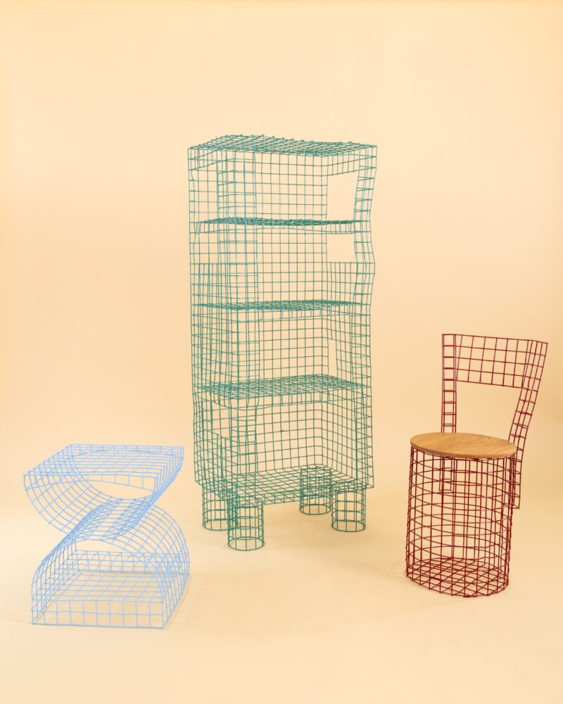 Four geometric furniture pieces made of colored wire mesh are arranged against a plain beige background, including a blue zigzag table, a green shelf, and a red chair with a wooden seat—each thoughtfully designed as part of sustainable home furnishings.