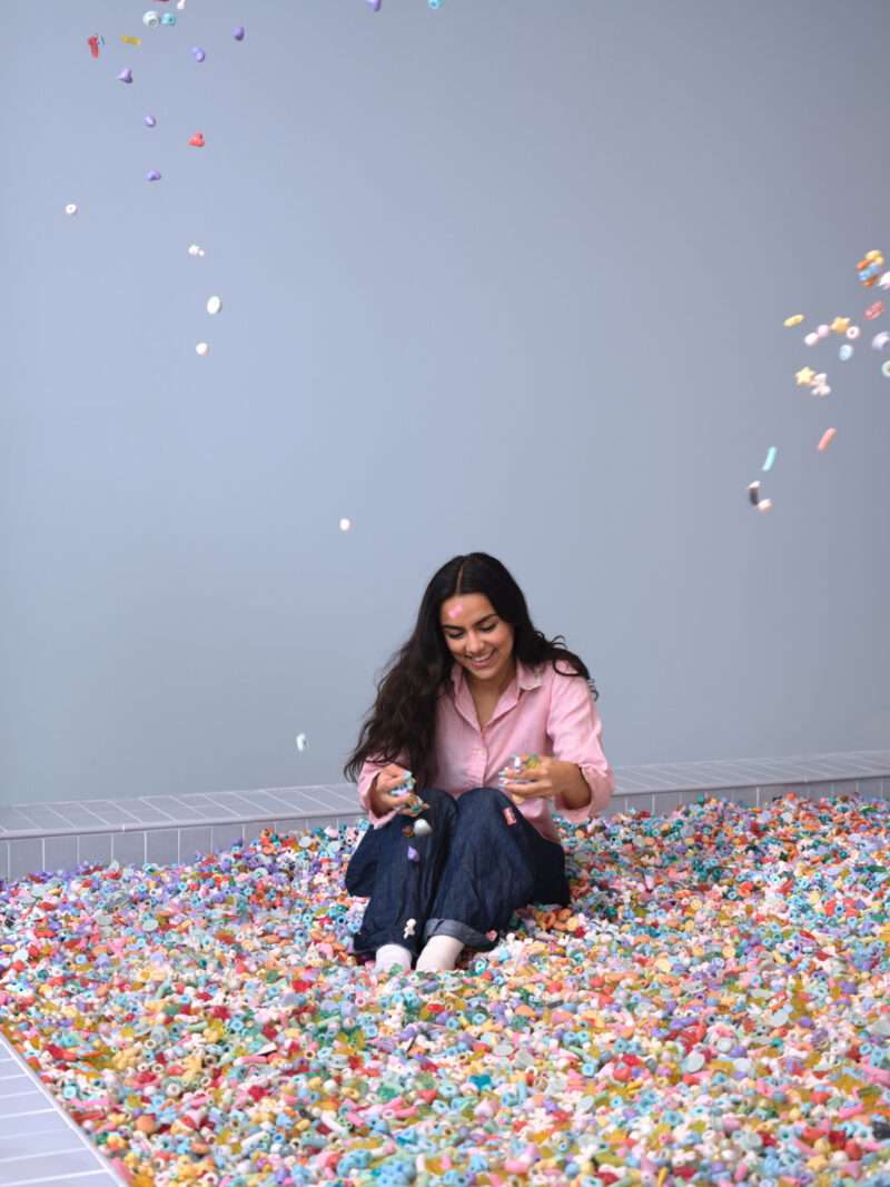 A woman sits on a floor covered with colorful small toys, smiling and holding toys in her hands while more toys fall around her against a plain light blue wall, evoking the playful atmosphere of a Cj Hendry Pastel exhibition.