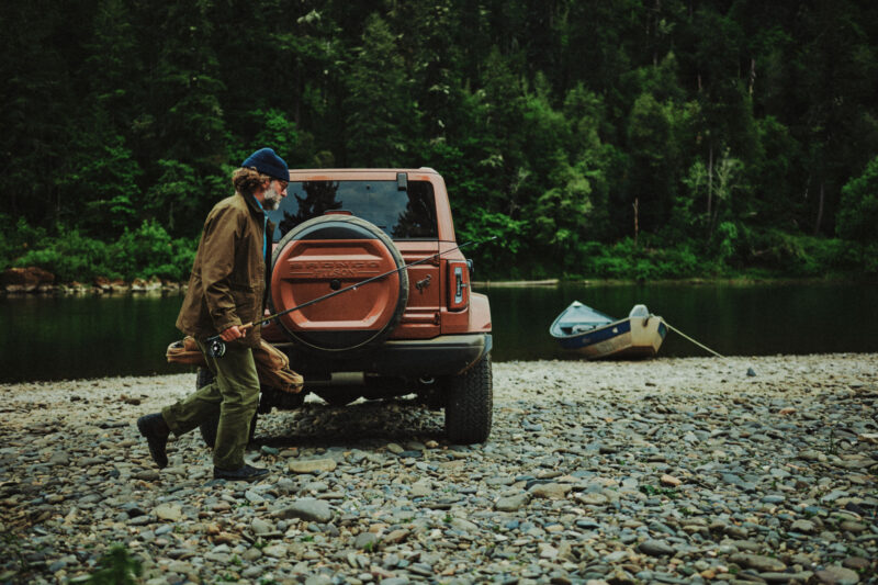 A person carrying a fishing rod walks past an SUV parked on a rocky riverbank, with a boat tied to the shore and a forest in the background.