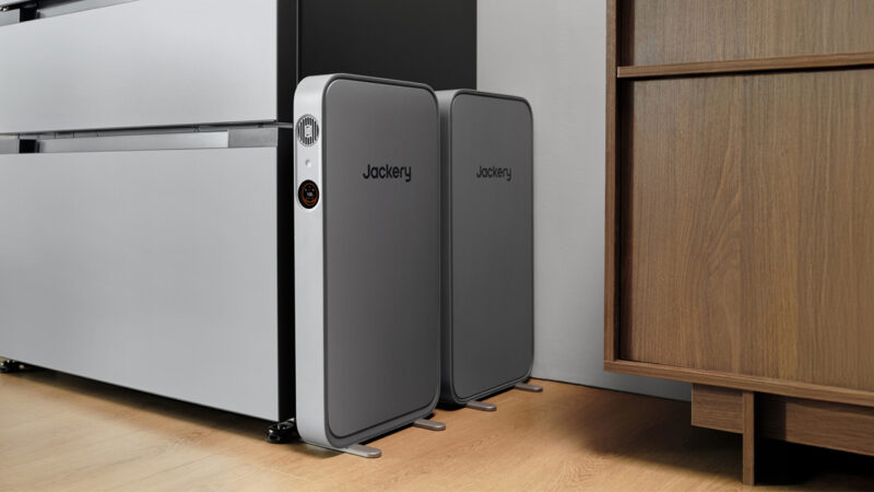 Two Jackery branded portable battery units are placed upright on a light wood floor between a black-white cabinet and a wooden dresser.