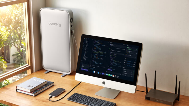 A desktop computer, keyboard, and router sit on a wooden desk near a window, with a large Jackery device placed upright beside them.