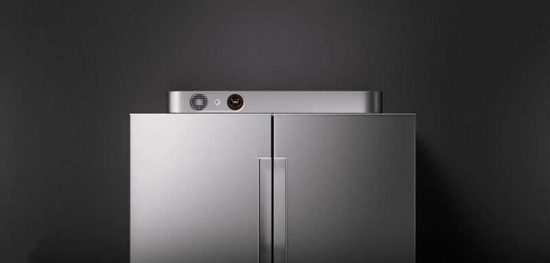 A metallic double-door refrigerator stands against a dark gray wall, featuring a small control panel and vent on the top edge.