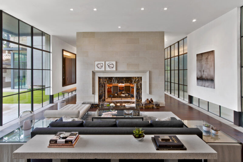 Modern living room with floor-to-ceiling windows, gray sectional sofa, marble fireplace, minimalist decor, and views of an outdoor lawn.