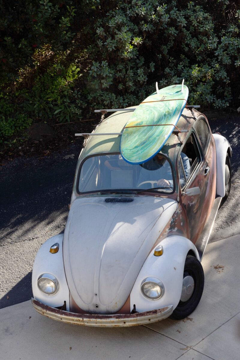 Vintage Volkswagen Beetle with worn paint parked on a driveway, carrying two surfboards on its roof.