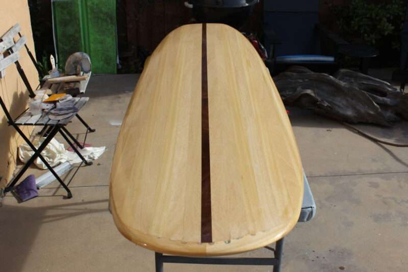 A wooden surfboard with a dark center stripe rests on sawhorses in an outdoor workspace, surrounded by tools and chairs.