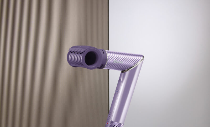 A close-up of a sleek, purple Shark FlexStyle IonCurl handheld hair styling tool with a hollow, cylindrical head, positioned against a minimalist background with textured and smooth surfaces.