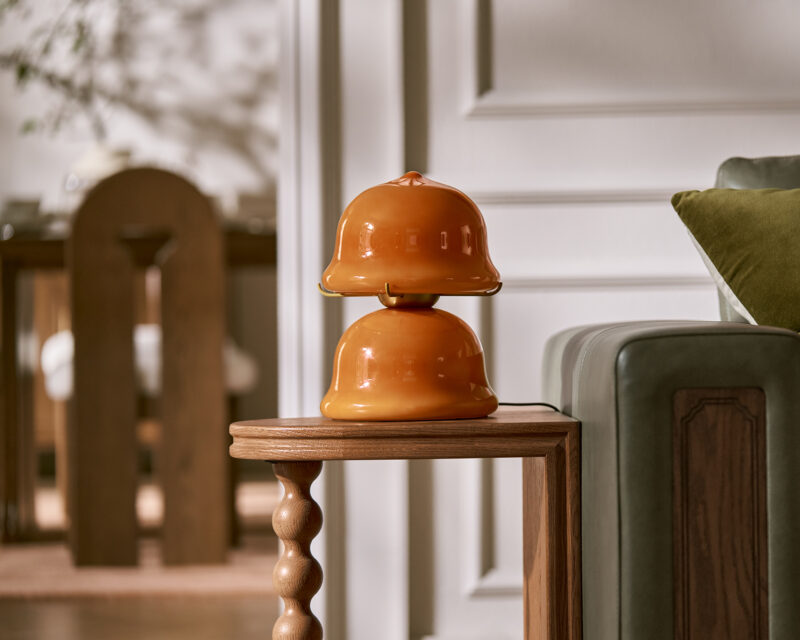 Two orange ceramic bell-shaped objects are stacked on a small wooden side table from the Tan France Castlery furniture collection, positioned next to a green sofa in a well-lit living room.