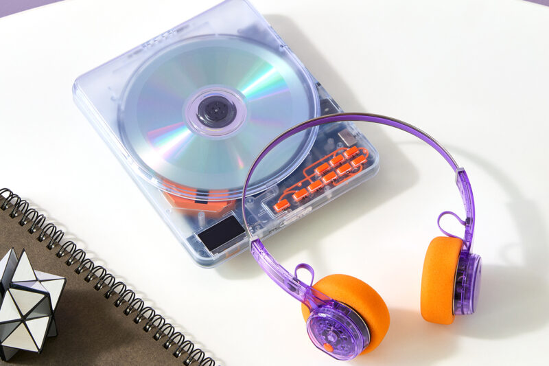 A portable CD player featuring a transparent retro tech design with a translucent cover, orange and purple headphones, a spiral notebook, a pen, and a geometric puzzle on a white table.