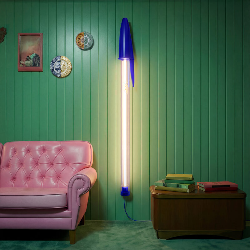 A tall lamp shaped like a blue pen, inspired by transparent retro tech, stands by a pink sofa, with a wooden side table and wall art on a green paneled wall.