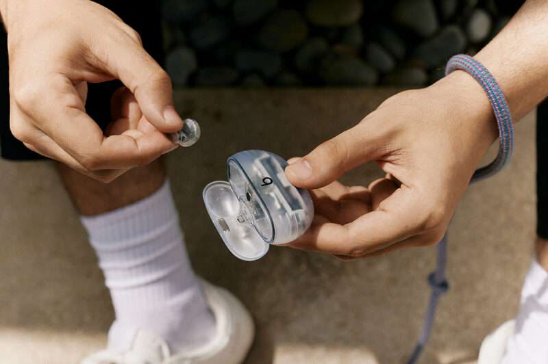 A person holds a small open container labeled “9” and places a gray earbud inside. The transparent retro tech vibe of the scene stands out, with the person wearing white socks, white shoes, black shorts, and a blue bracelet.