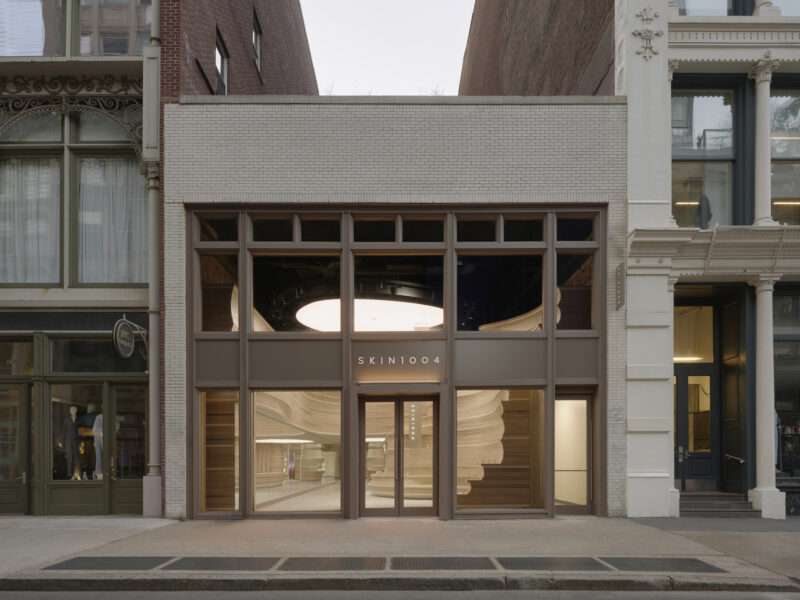 Storefront of SKIN1004 with glass doors and windows, revealing a modern interior where a prominent spiral staircase rises beside neutral-toned walls—echoing the aesthetic that defines SKIN1004?s first NYC store, milled from stacked wood.
