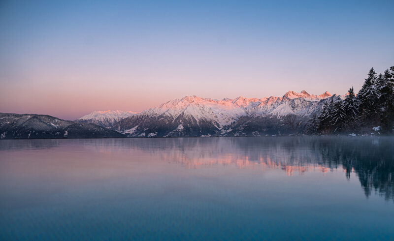 An infinity pool overlooks snow-covered trees and distant mountains at sunrise, with the peaks reflecting pink hues from the early morning light.
