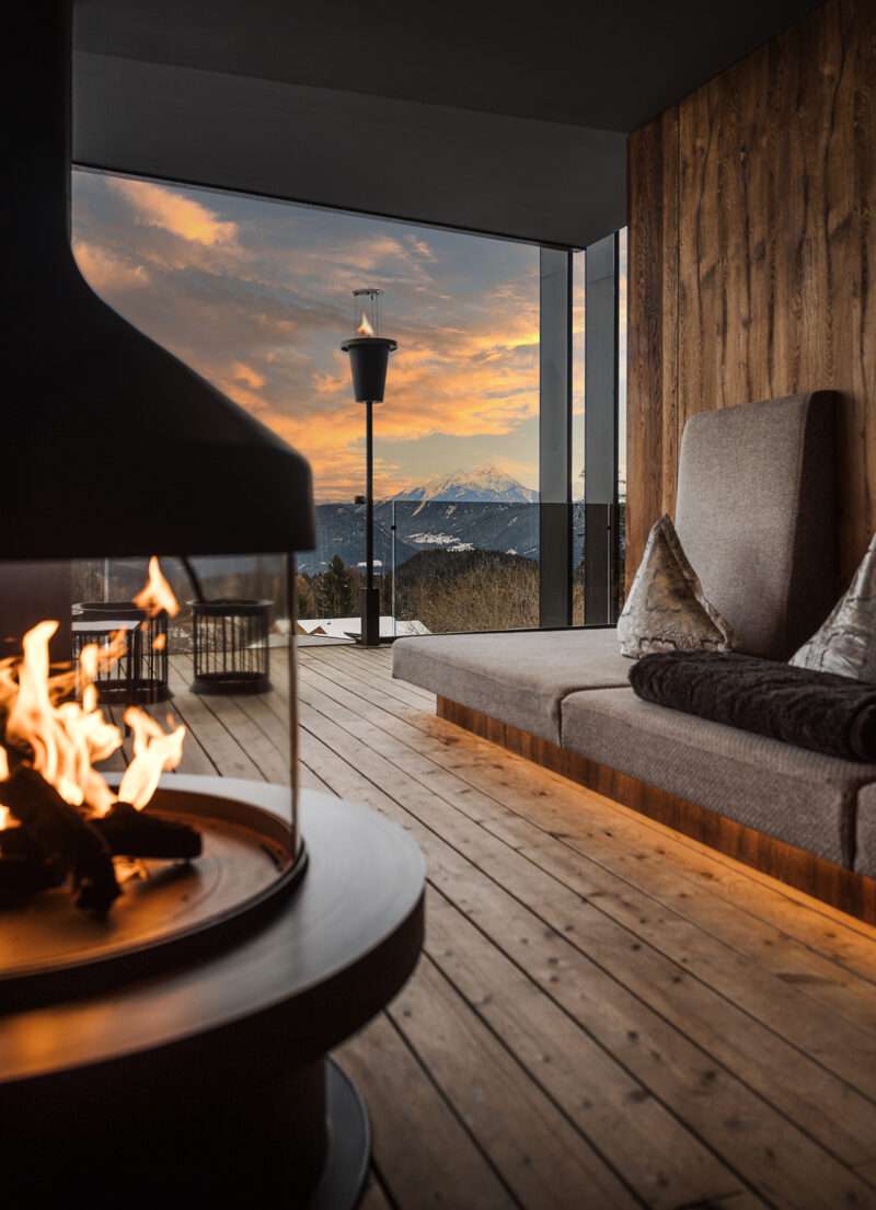 Modern living room with a round fireplace, wooden floor, and large windows offering a mountain view at sunset, reminiscent of the refined elegance found at Hotel Chalet Mirabell.