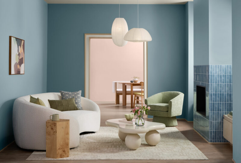 Modern living room with Valspar 2027 Color of the Year blue walls, a white curved sofa, green chair, abstract art, two pendant lights, and a round table with flowers; a dining area is visible through an open doorway.