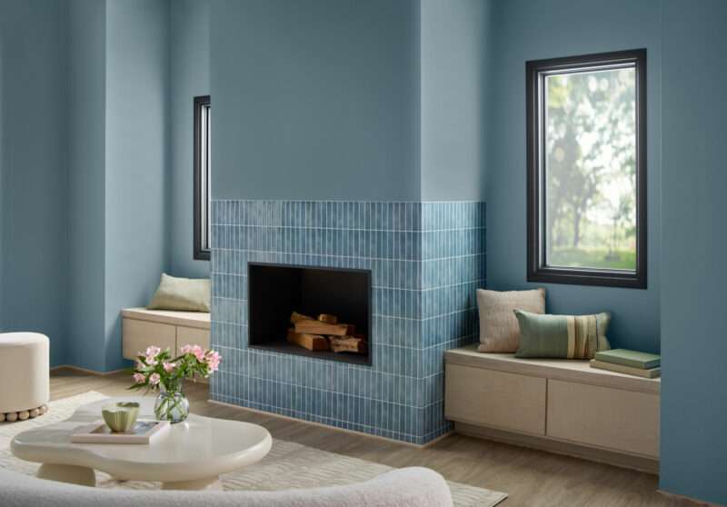 A living room with a blue-tiled fireplace, built-in window seats with cushions, blue walls painted in Valspar 2027 Color of the Year, wood flooring, and a round coffee table with a vase of flowers.