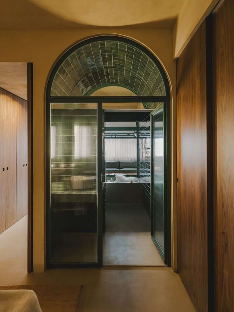 A glass door with a green frame and arched window opens to a room with bunk beds. Walls are wood-paneled; the floor is beige. The scene is softly lit with natural light.