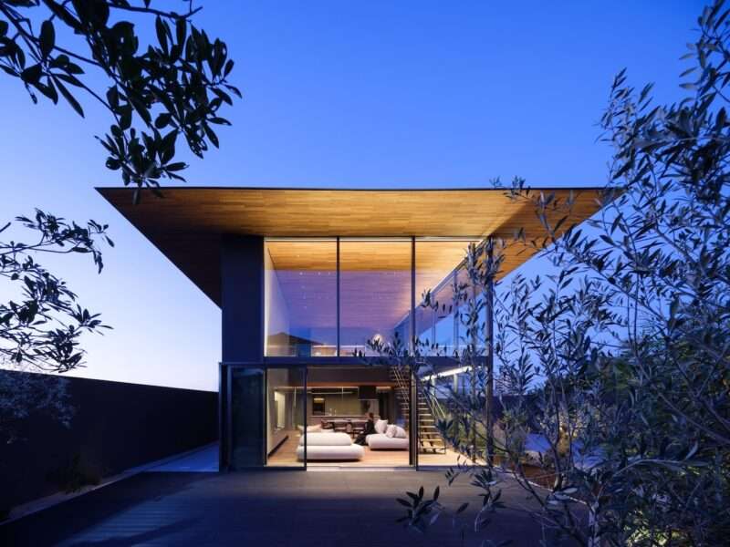 Modern house with large glass walls and a flat wooden roof, illuminated interior visible at dusk, surrounded by trees.