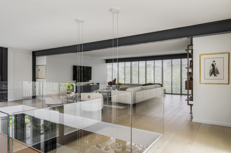 Modern living room inspired by the Paul Rudolph Biggs Residence, featuring a white sectional sofa, TV, bookshelves, large windows with blinds, wood floor, and glass railing over a staircase.