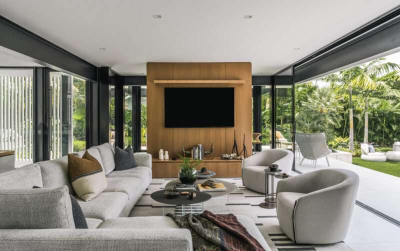 Modern living room with gray sofas, round chairs, a wood-paneled TV wall, and large windows overlooking a lush garden, inspired by the Paul Rudolph Biggs Residence.