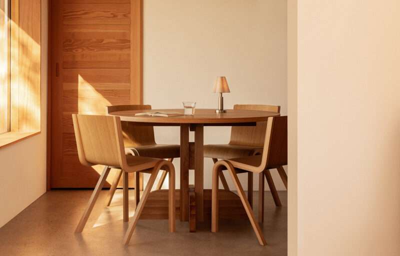 A round wooden table with four matching chairs and a small lamp sits in a sunlit room with wooden walls and a glass of water on the table.