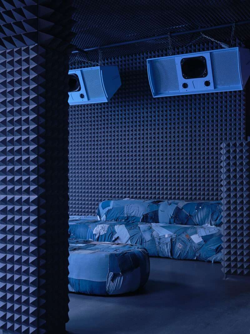 Room with foam-padded walls, blue upholstered seating, and two large retro TVs hanging from the ceiling. The scene has a blue lighting ambiance, perfectly capturing the modern yet nostalgic aesthetic of a Clap Studio design.