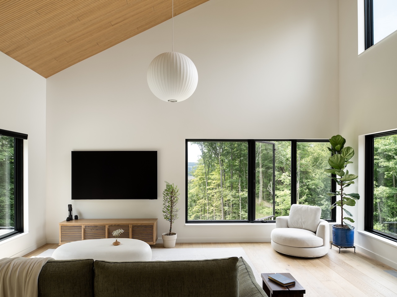 Modern living room with large windows, wall-mounted TV, green sofa, round white chair, potted plants, and light wood ceiling. Natural light fills the space.