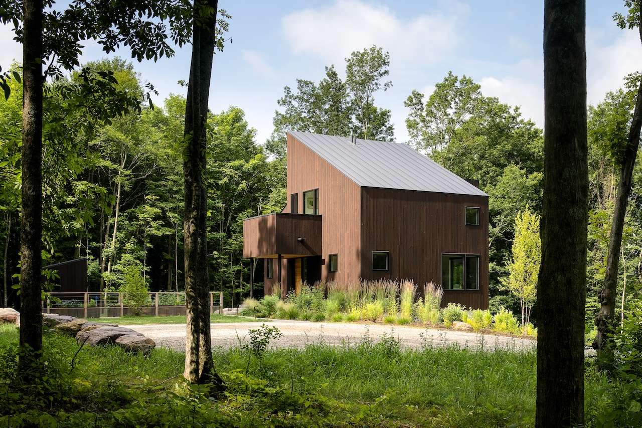 Modern two-story house with dark wood siding and metal roof, surrounded by trees and greenery, with a gravel driveway in front.